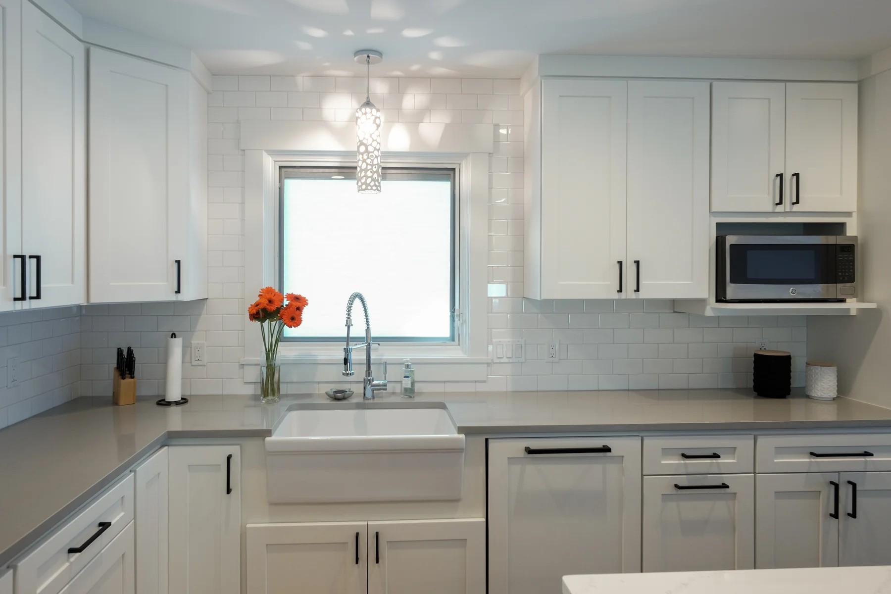 Baptist Lake kitchen — all-white shaker cabinets, white subway tile, gray quartz counters, a farmhouse sink, and black hardware, with orange flowers on the sill.