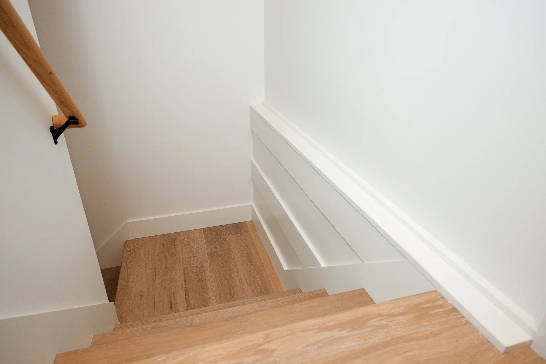A stairwell looking down — white walls, white-oak treads, and an oak handrail on black brackets.