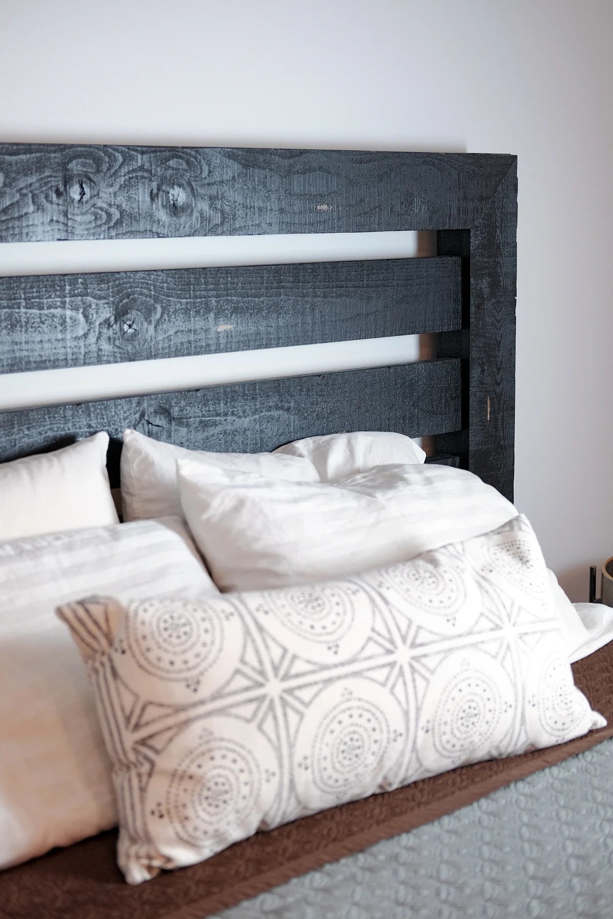 A bedroom detail with a black-stained reclaimed-wood pallet headboard, white linens, and a patterned lumbar pillow.