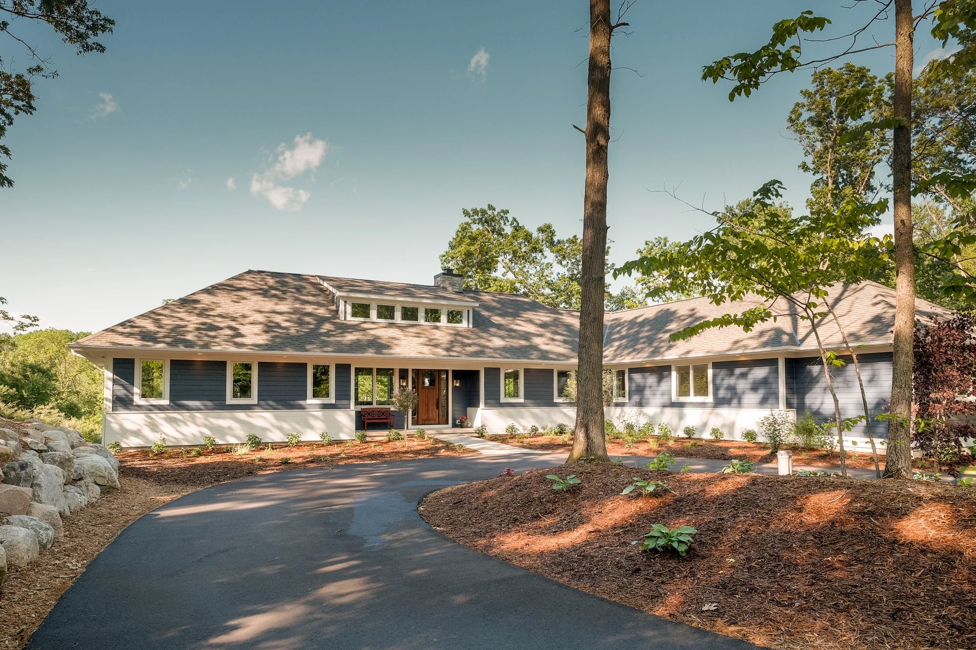 Fairbanks Estates front exterior — gray-navy siding, cedar-tone roof, gabled center entry, curved drive through a mature wooded lot under a blue sky.
