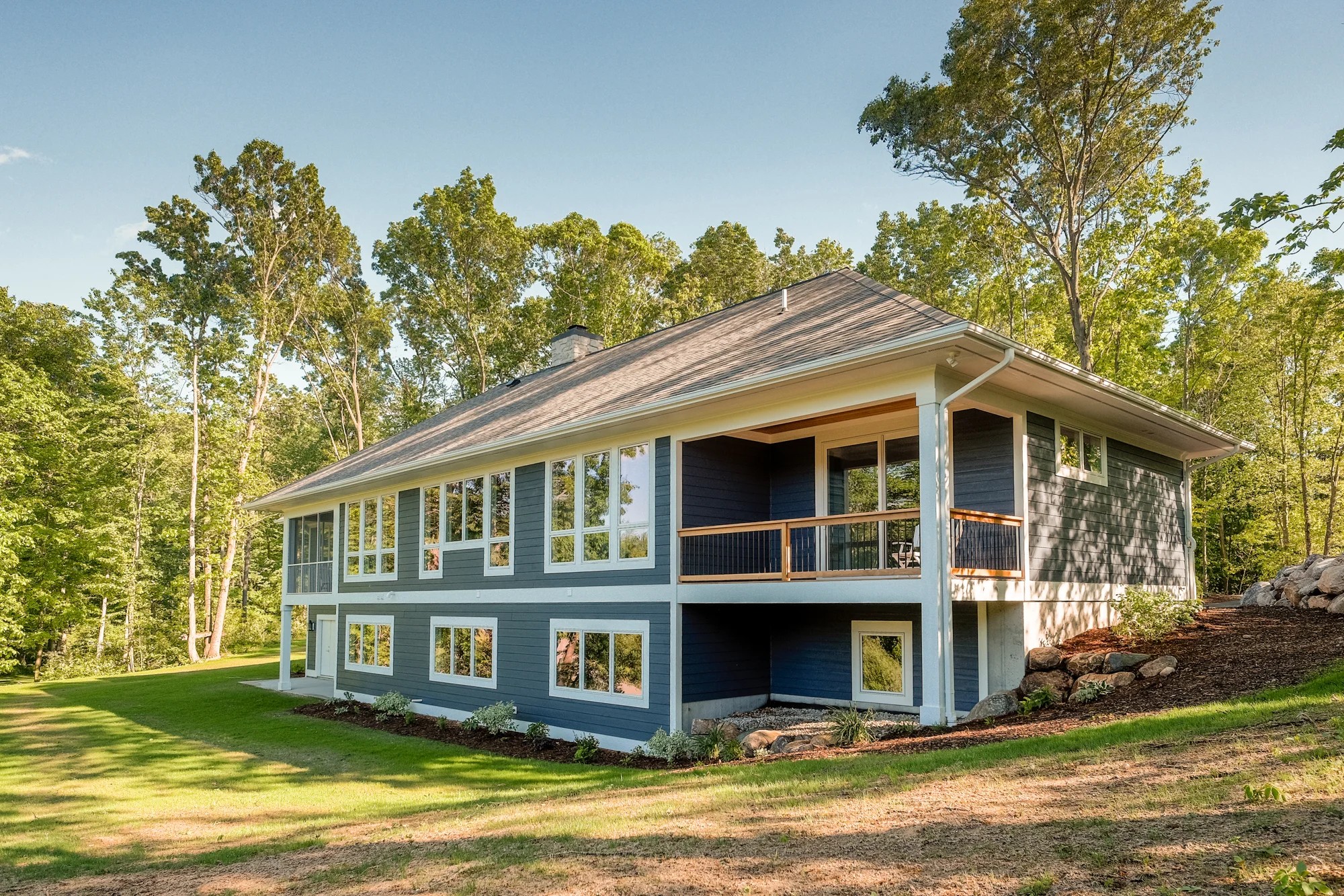 Fairbanks Estates rear and side exterior — navy siding, a covered upper deck, a walkout lower level, and a green sloped lawn against the woods.