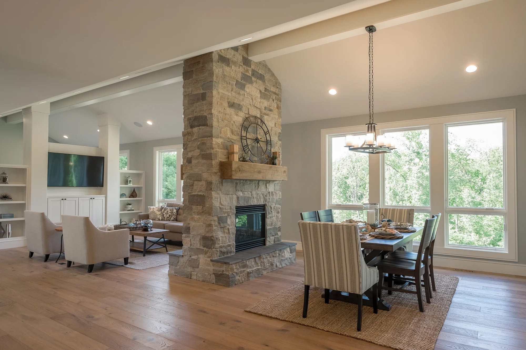 Open dining room and great room joined by a double-sided stacked-stone fireplace with a reclaimed-wood mantel and a wall of windows looking into the woods.