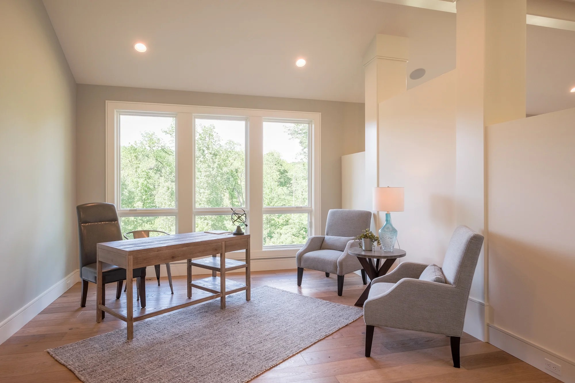 A bright home office and sitting room with a large window wall to the woods, a wood desk, and gray chairs.